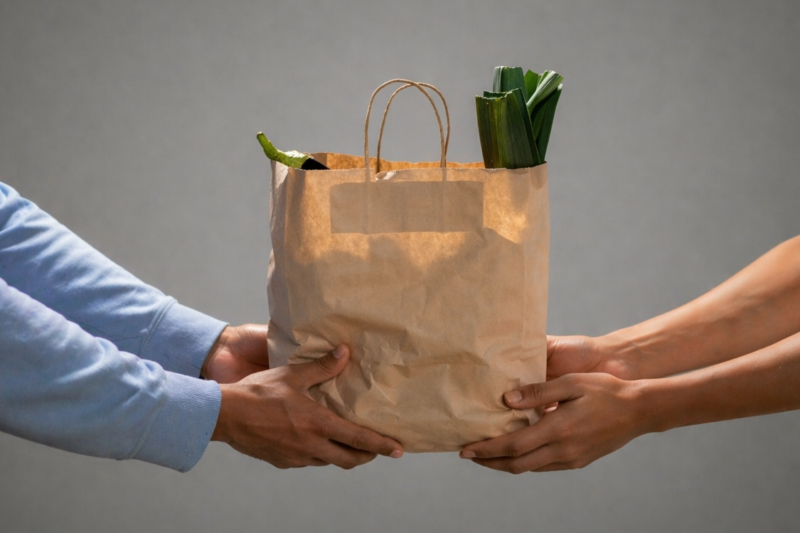 Person handing over a brown paper bag with groceries to another person against a gray background
