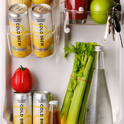 Inside of a brightly lit refrigerator door featuring four cans of Pilot Coffee Roasters Black Cold Brew stored alongside everyday items including a red tomato, a bunch of celery, a glass water bottle, a green apple, and a hanging set of keys.