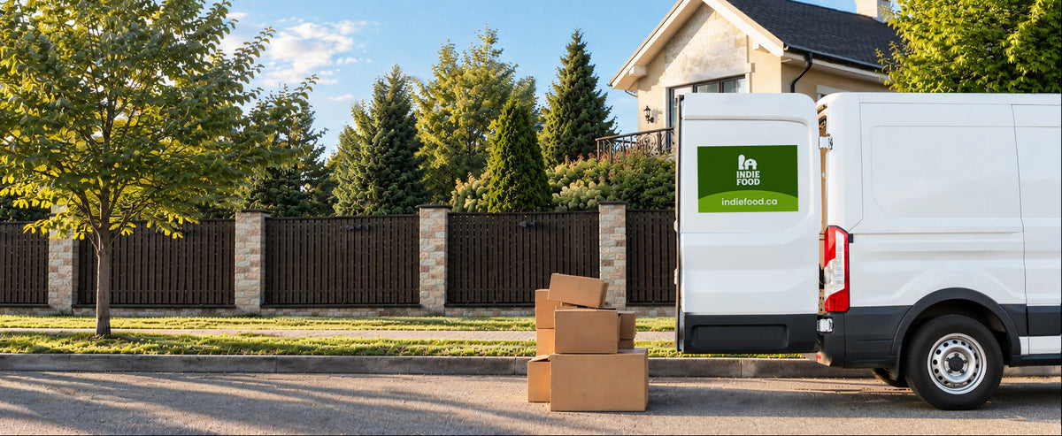 A white moving truck parked on a suburban street with its back door open, next to stacked cardboard boxes. The truck has a green logo on the side door.