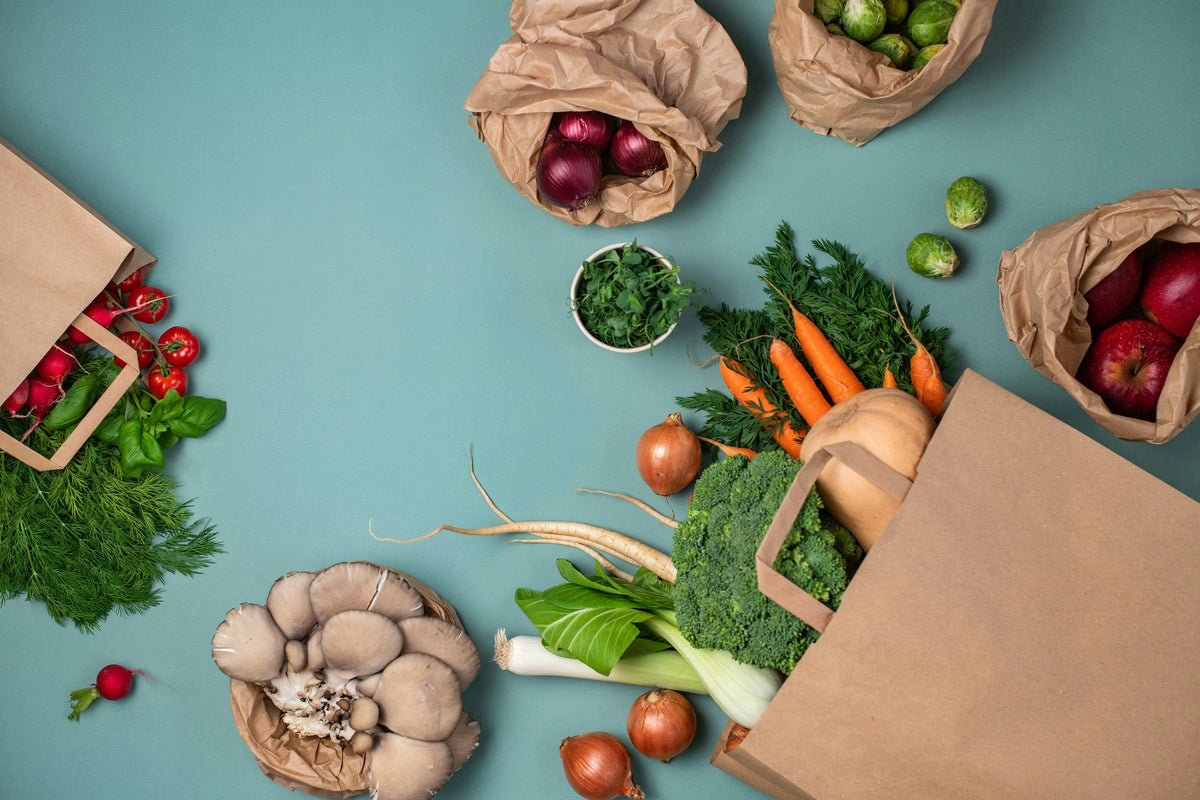 A styled flat-lay of fresh groceries in brown paper shopping bags on a teal background, including broccoli, carrots, apples, mushrooms, radishes, and herbs.