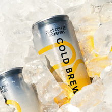 A refreshing close-up of a silver and yellow Pilot Coffee Roasters Cold Brew can nestled deeply in a bed of crushed ice under bright, sunny lighting.